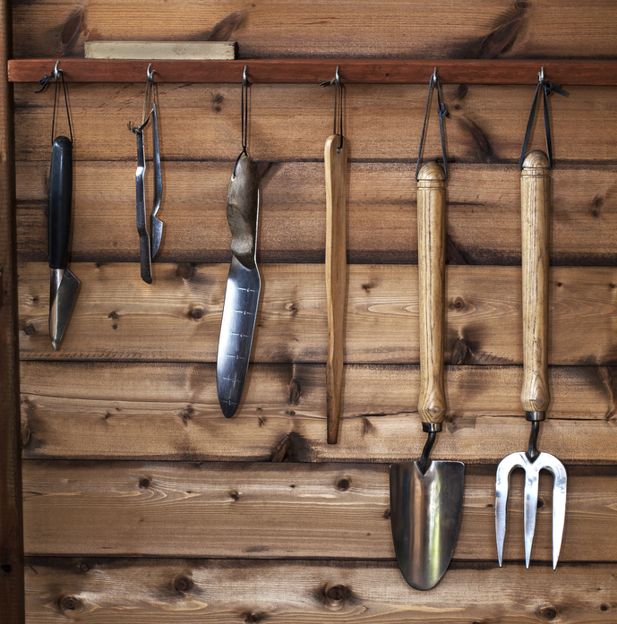 Gardening tools hanging on a wooden wall, including a trowel and fork.