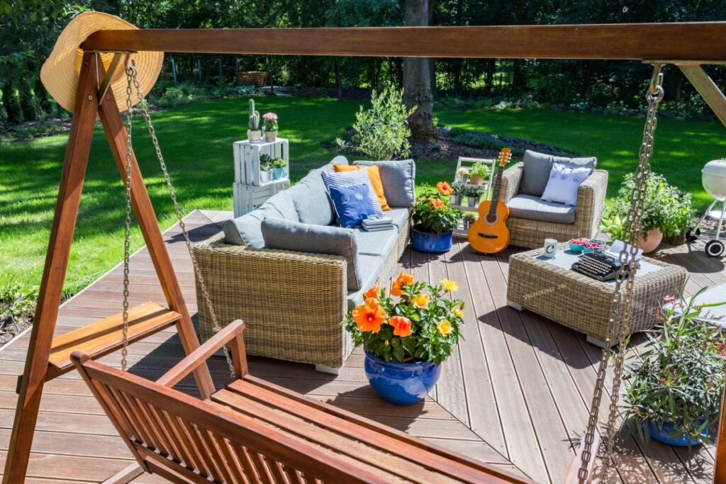 Cozy garden lounge set with swing, wicker furniture, cushions, and potted flowers on a wooden deck.
