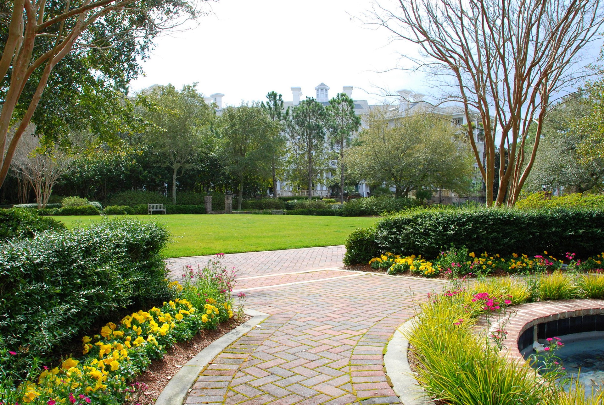 Brick garden path leading to a lawn, trees, and a building in the background.
